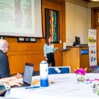 two people watching a woman present at a podium in a conference room